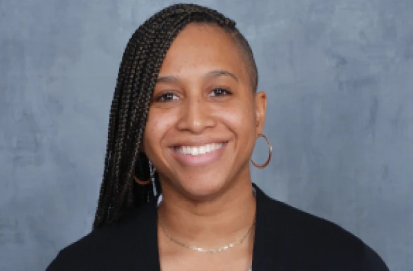 Smiling woman with braided hair, wearing hoop earrings and a black top, posing against a gray background.